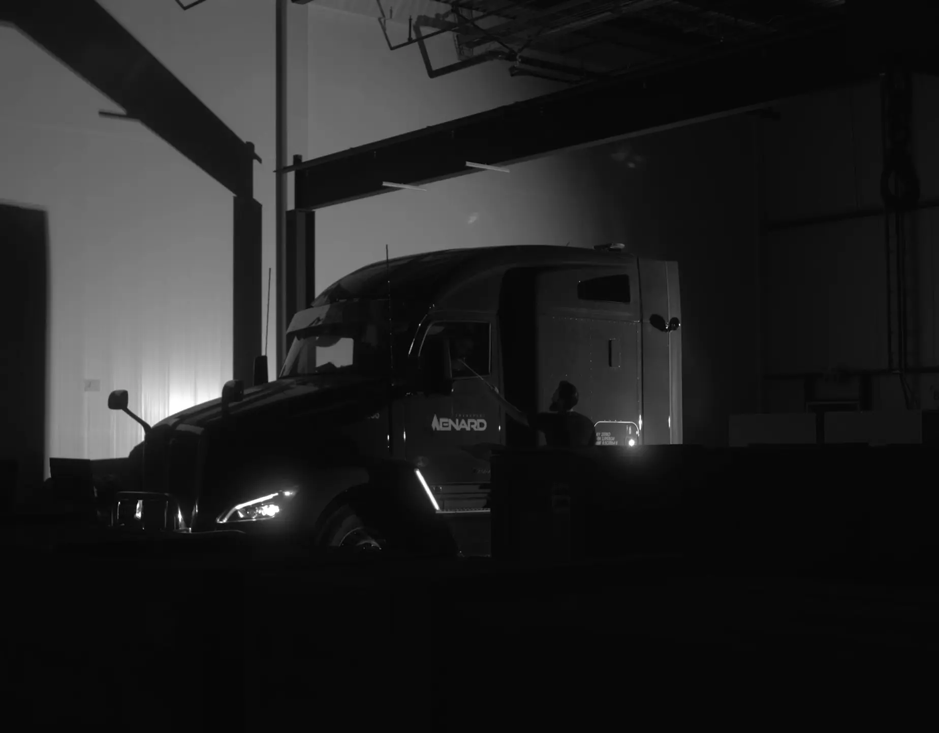 Dim indoor loading bay with a large semi-truck parked inside; headlights glow on the front bumper, backlit by a faint wall light. The truck bears a visible company name on its door.
