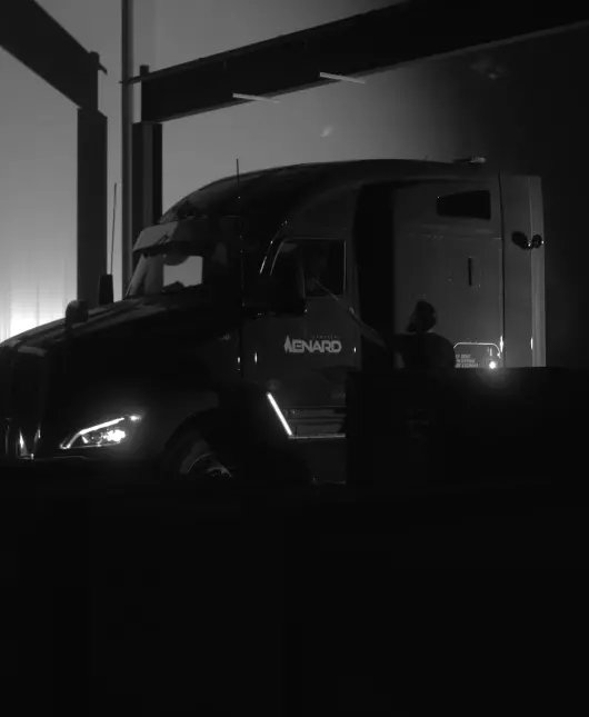 Dark nighttime photo of a large semi-truck cab at a loading dock, headlights on and driver inside, logo on the door barely visible.